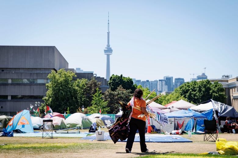 A person carrying banners and posters walking on the grounds of an encampment littered with tents at UofT and the CN Tower in the distance 