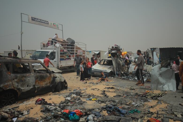 Burned out vehicles and large amounts of debris and clothing are shown on a sandy ground, with adults and children milling around.   