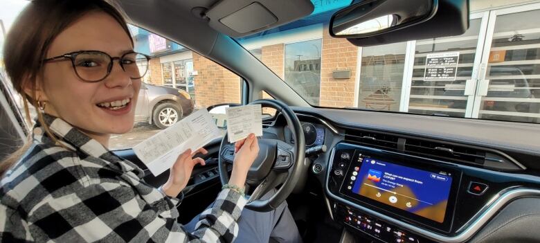 A blond girl wearing glasses and in the driver's seat of a car shows off two pieces of paper.