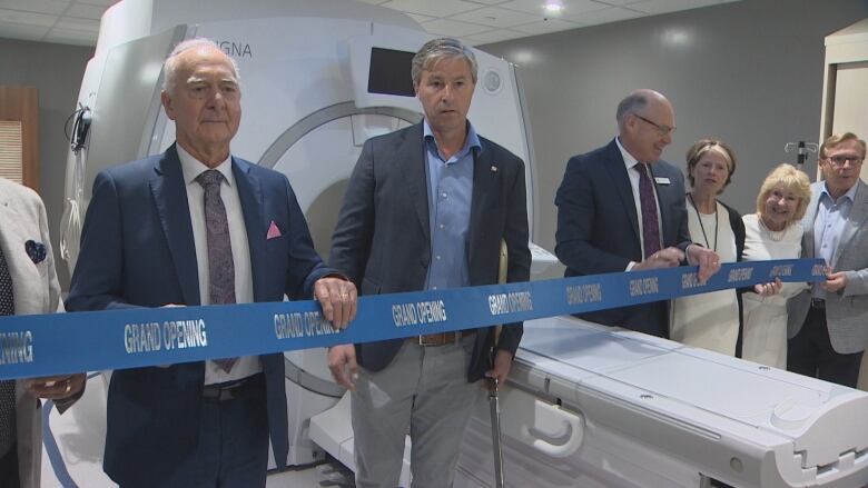 A group of people, one of whom has a giant pair of scissors, stands behind a blue ribbon strung in front of an MRI machine