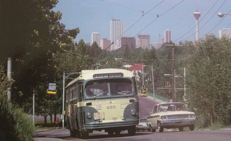 A trolley bus drives on a road with a city skyline behind it.