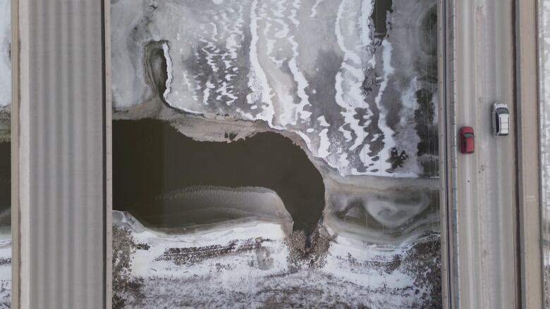 An overhead shot shows two spans of a bridge crossing a river. Part of the river is frozen, while an area spreading out from a culvert is clear of ice