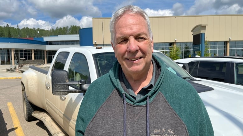 A white man wearing a green-tinged grey hoodie smiles while in a parking lot.