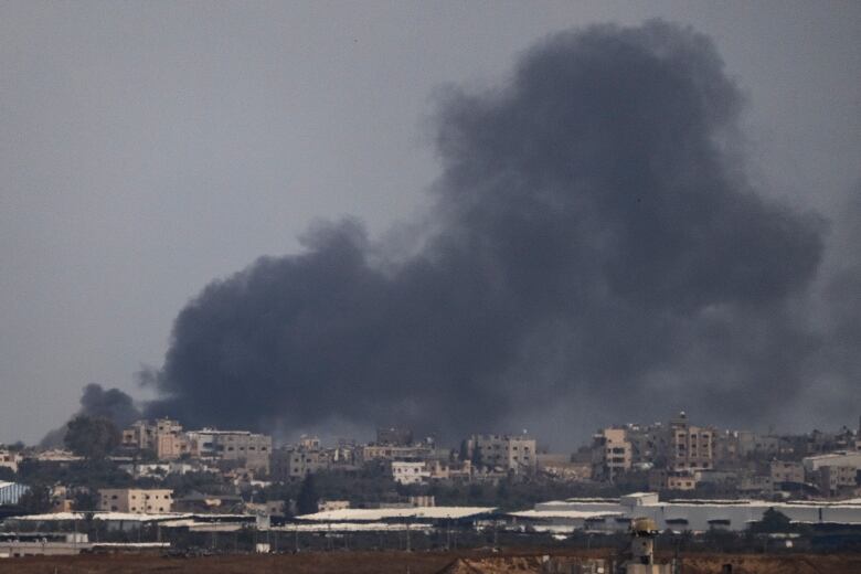 A large cloud of dark smoke is shown in the distance about a city skyline that consists of low-rise buildings.