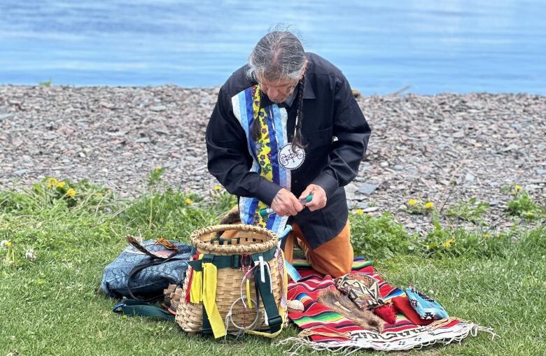 A man with grey hair and wearing a colourful shirt bends down over a woven basket and colourful objects strewn over green grass. A river flows behind him.