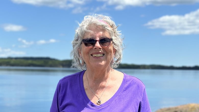 A smiling woman with white, curly hair and wearing sunglasses and a purple shirt and skirt stands in front of a river.