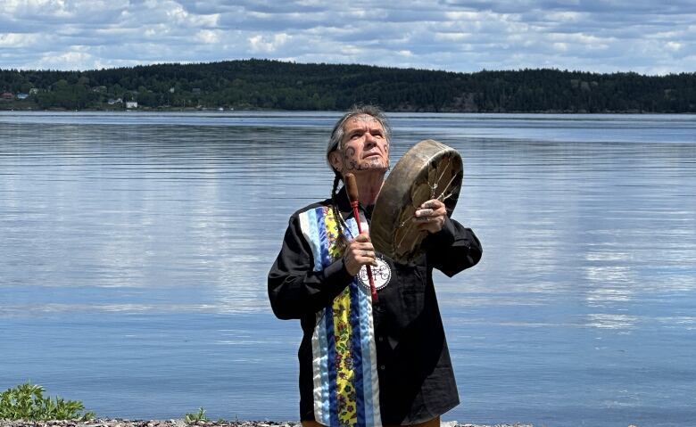 A man with grey hair braided down his back stands in front of a flowing river, banging a hand drum and looking up to the sky.