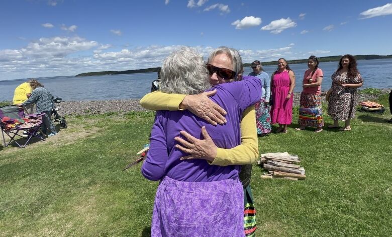 A woman wearing sunglasses and a yellow shirt stands outside on green grass in front of a river hugging another woman wearing a purple shirt and skirt.