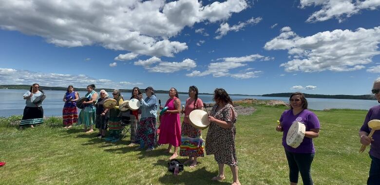 A row of women wearing colourful clothing stand outside on green grass holding up hand drums. A river flows behind them.