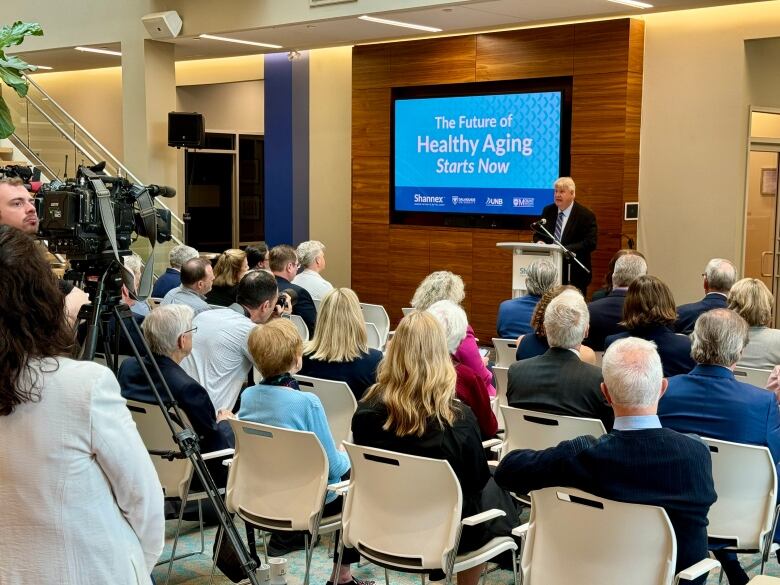 Man in suit stands in front of microphone in front of a room full of people.