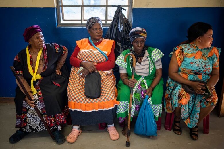 Four women are shown seated in a row, with three of them wearing headdresses and all of them wearing multicolored dresses.