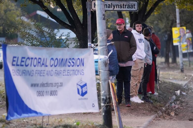 A handful of people are shown outdoors standing under a street sign as a nearby sign reads, 'Electoral Commission: Ensuring Free and Fair Elections.'