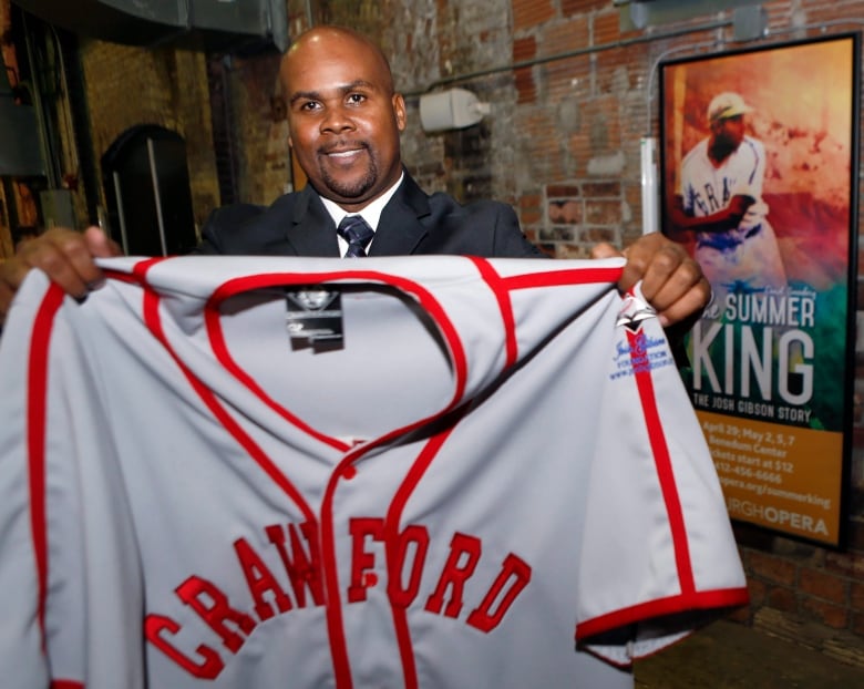 A balding man with a beard holds up a baseball jersey that says 'Crawford.'