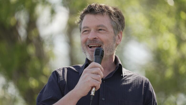 A man in a dark collared shirt holds a microphone and stands in front of trees.