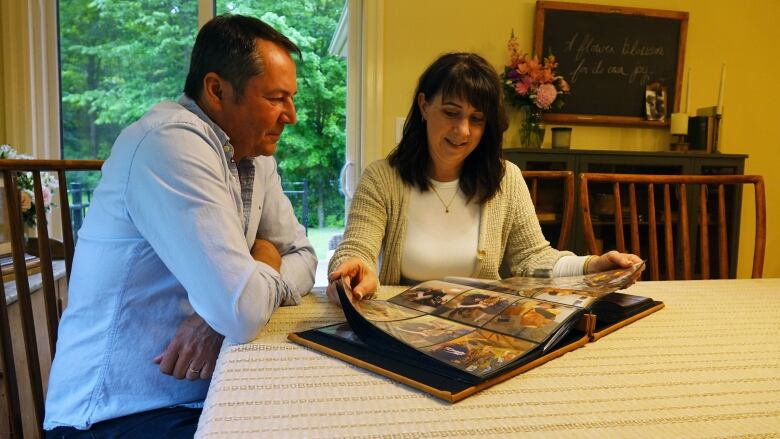 A man in a blue shirt and a woman with long dark hair smile at a photo album. They're sitting at the kitchen table.