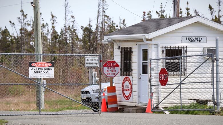 A small white building sits behind a chain-link fence covered with black, red and white warning signs saying danger active mine and stop proceed only when directed.