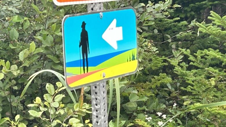 A sign depicting a hiker and an arrow indicating left. Bushes and a tree in the background.