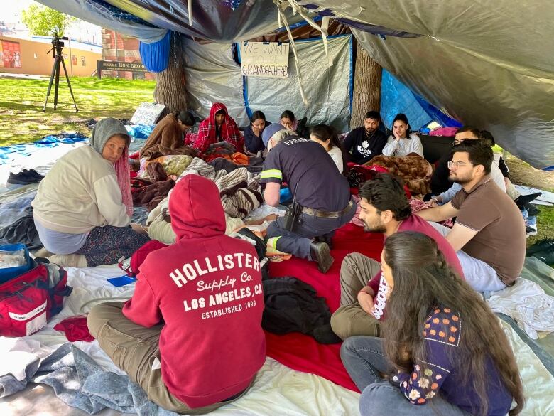 A group of people sit around the ground in an encampment as a paramedic tends to one of the people.