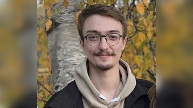 A man with short hair and glasses smiles for a photo outside in front of a birch tree.