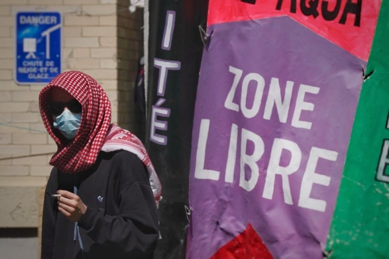 white person wearing a red kuffiyeh, a surgical mask and black hoodie smokes a cigarette next to a purple poster that says 