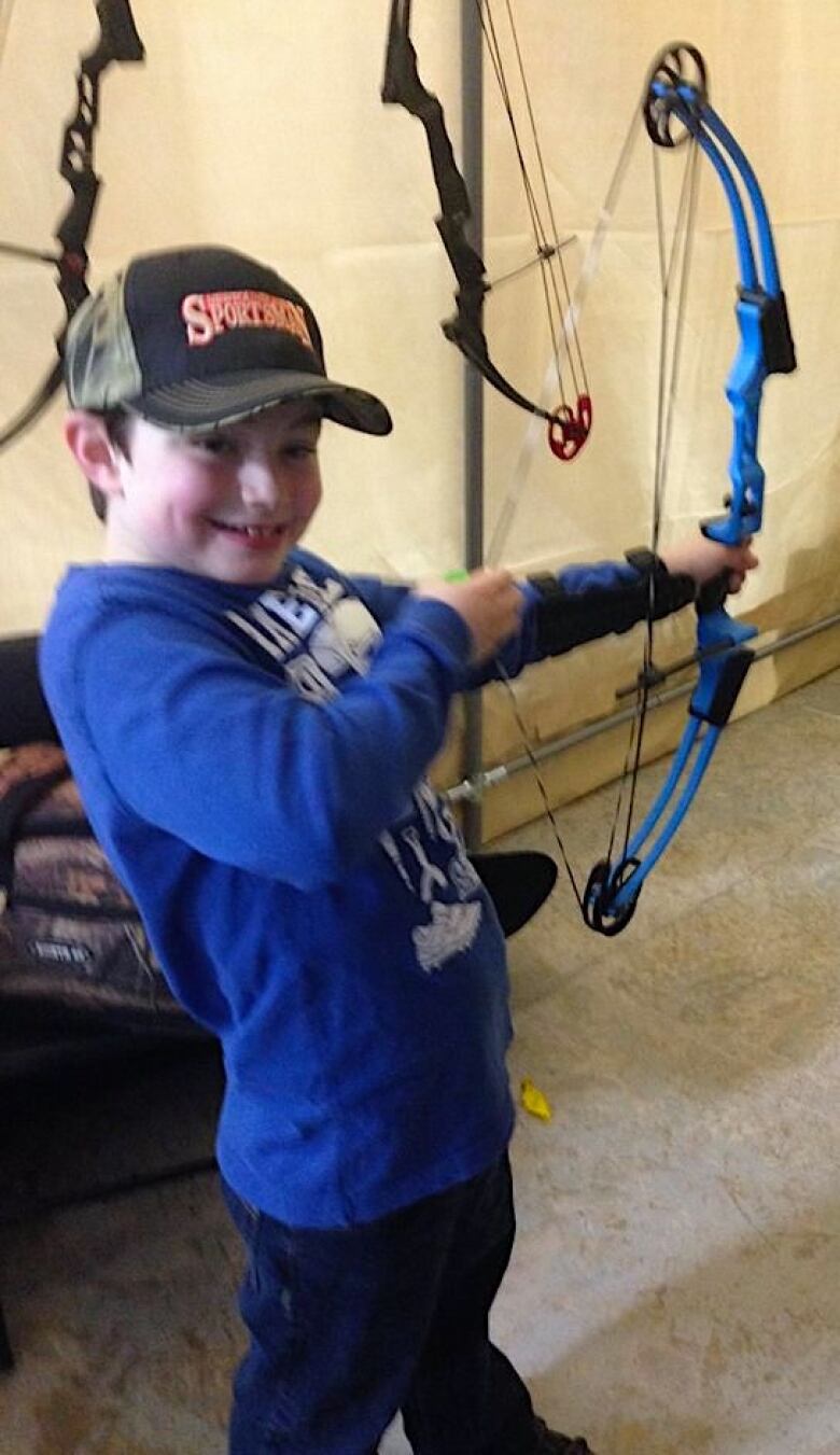 A young boy in a baseball cap and a blue shirt pulls back the string on a bow.