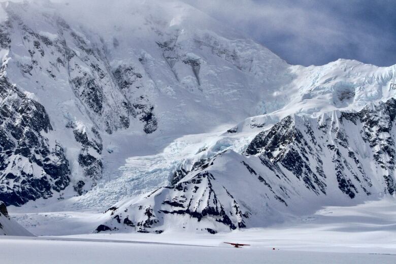 A small plane is seen from afar on the snow near a rugged mountain peak.