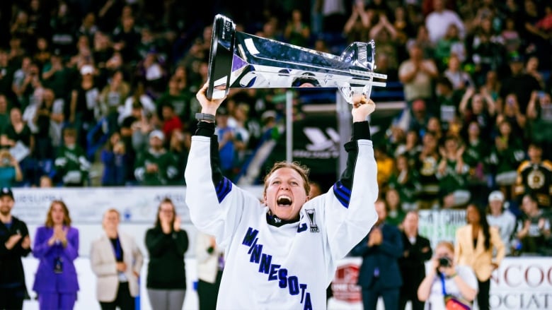 A woman wearing a white Minnesota hockey jersey lifts a silver championship trophy above her head.