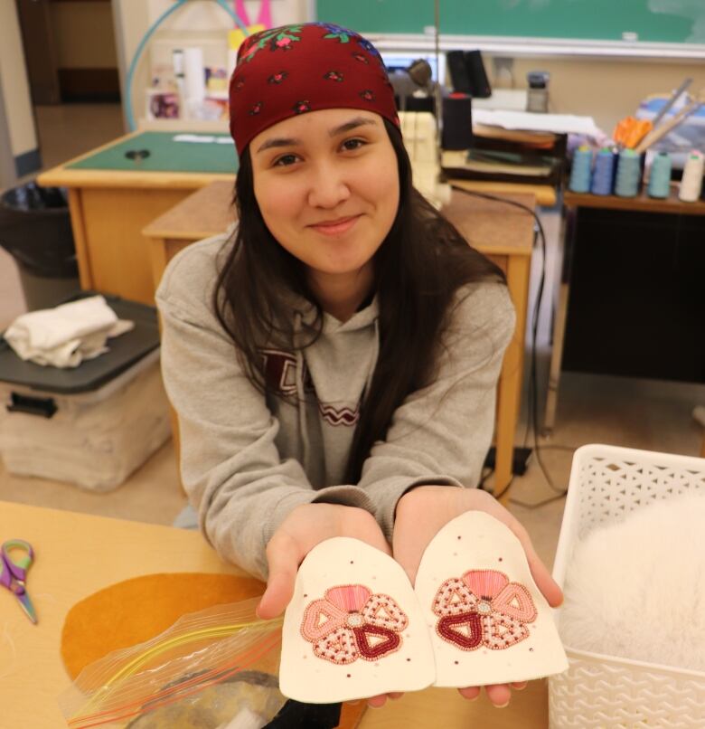 A young woman sits at a table holding up some beaded moccasin tops.