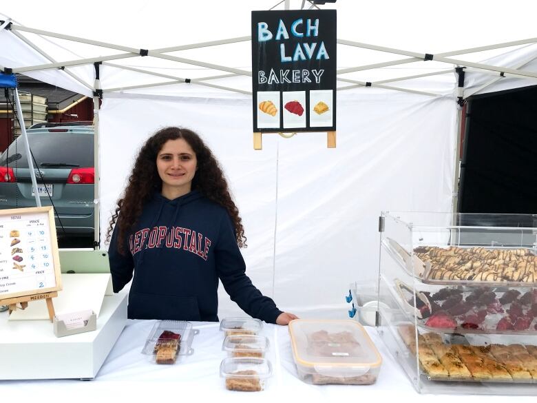 A young woman with long curly hair stands in front of a baklava stand at a farmer's market.