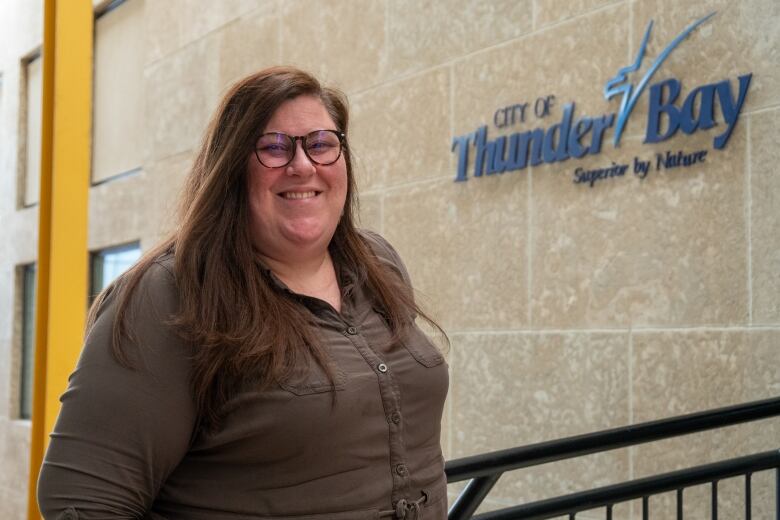 A person with long brown hair and glasses stands in front of a sign that says 