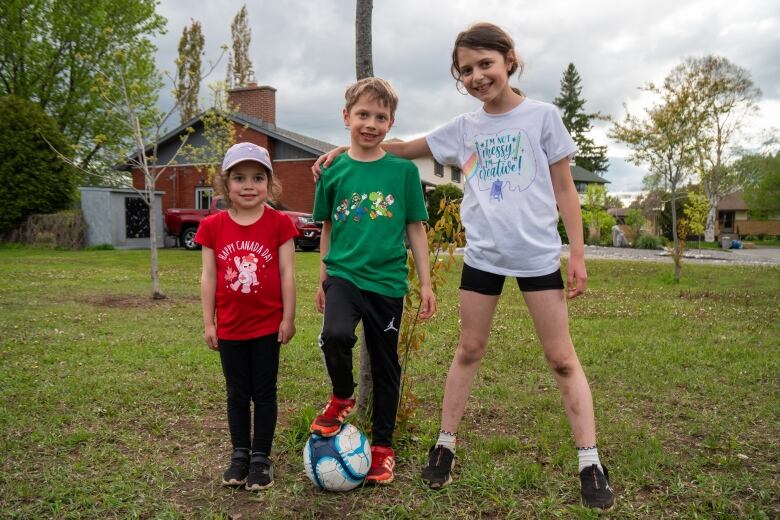 Three children stand in a green space, smiling. The child in the centre has their foot on a soccer ball.