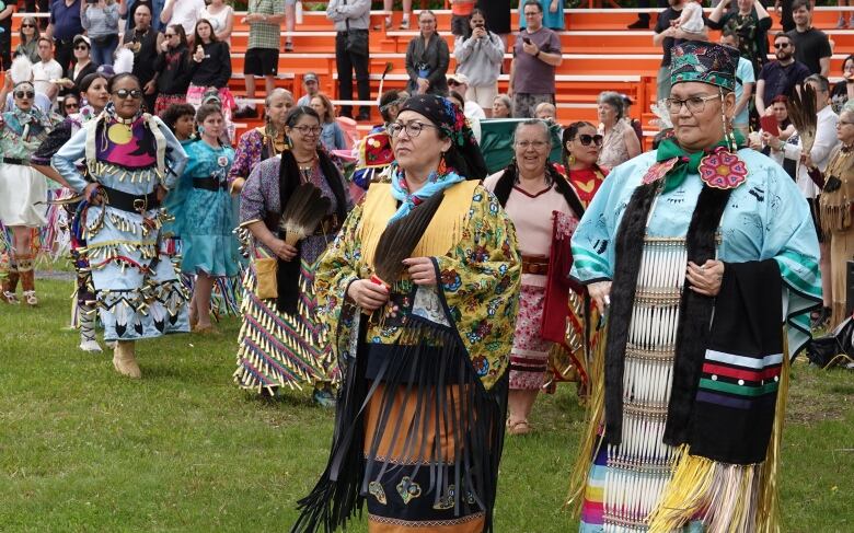 Indigenous women in regalia grouped together, walking across a grass field. Two older women are leading the group, one wearing blue tones and the other wearing yellow tones. People in stands are watching them in the background.