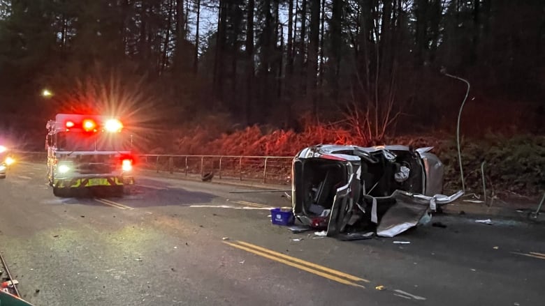 A flashing fire truck next to a destroyed and flipped over car on a road.