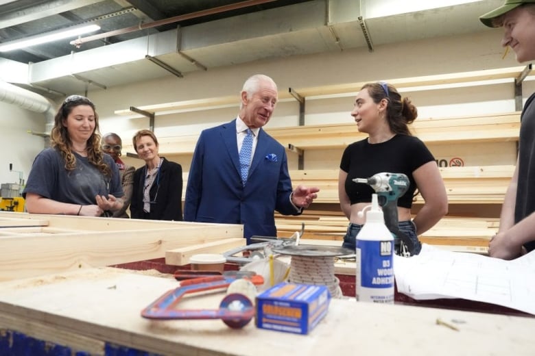 A person standing behind a table with pieces of wood on it talks to other people in a scenic art studio of a theatre.