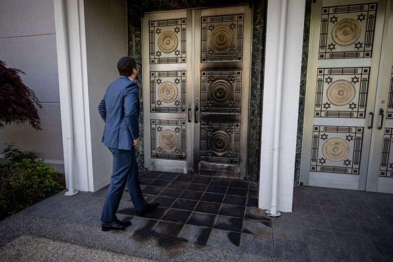 A man in a blue suit stands facing the doors of a synagogue, the doors have significant black burn marks on them