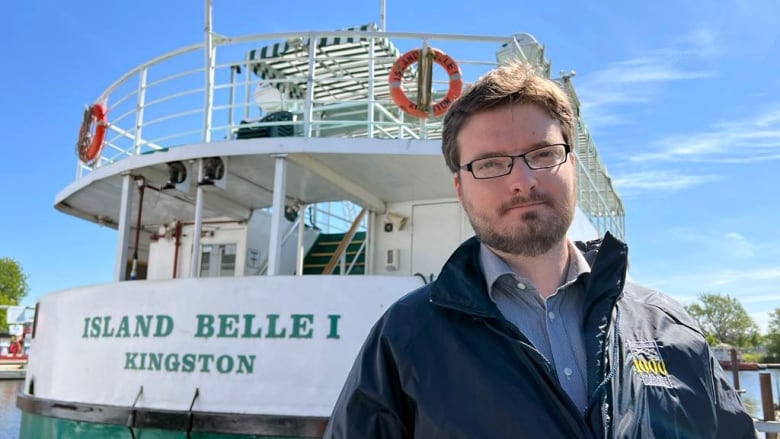 A man with glasses and a blue windbreaker stands in front of a large green and white passenger boat.