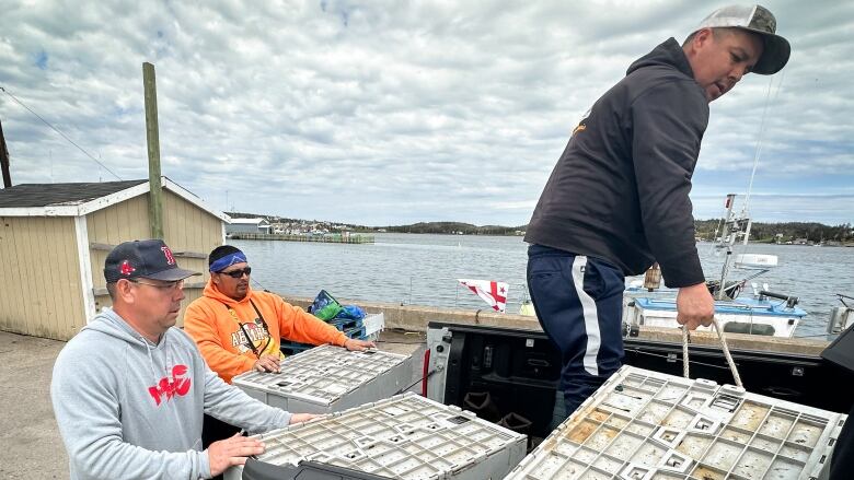 Two men push large plastic crates into the back of a pickup truck while a third man moves one by its rope handle, with a harbour in the background.