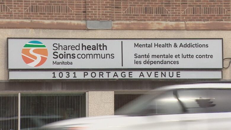 A white vehicle drives down the road, in front of an exterior sign for Shared Health's offices for mental health and addictions supports. 