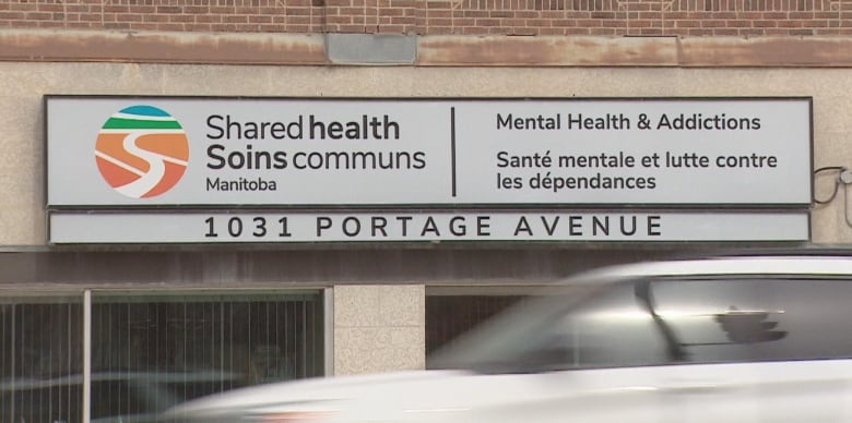 A white vehicle drives down the road, in front of an exterior sign for Shared Health's offices for mental health and addictions supports. 