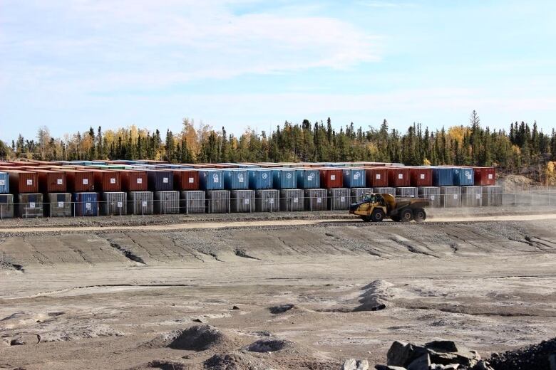 Shot from a distance away, a dump truck passes by a big row of different coloured sea cans.