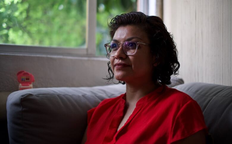 A dark-haired woman, wearing glasses and a red blouse, sits on a couch near a window.