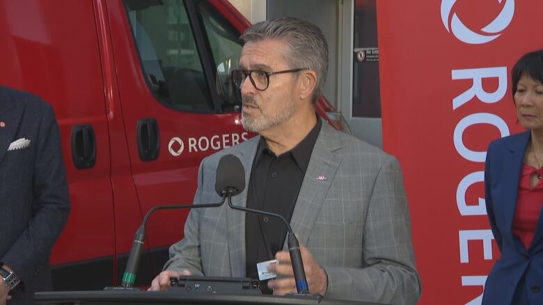 A middle aged man in glasses stands at a podium speaking into a microphone. The mayor of Toronto stands behind him