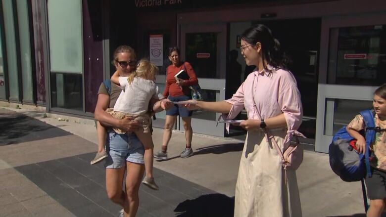 A young woman passes a flyer to a woman carrying a baby outside a city transit station on a warm spring day