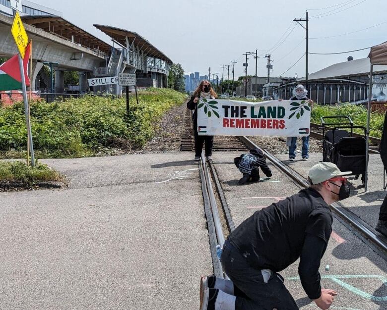 Two people holding a white flag between them that says The Land Remembers in black and red block a rail line. Another person kneels on the railway tracks.