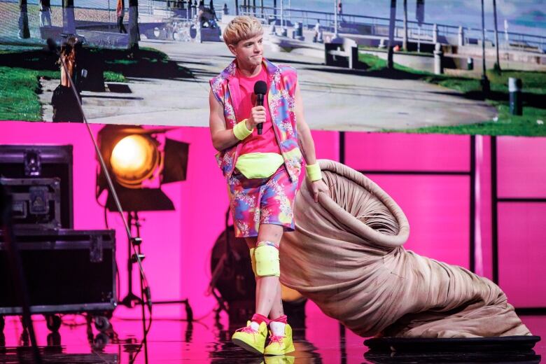 A person wearing a pink, purple, yellow and blue camo outfit stands onstage, holding a mic. They are dragging a large beige worm costume behind them. 