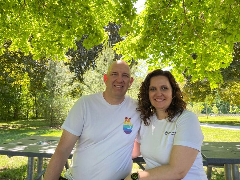 Two people sit on a bench under trees smiling. 