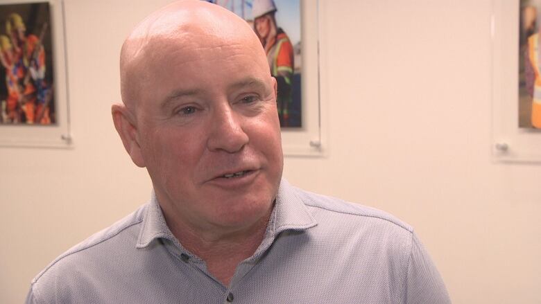 A bald man in a grey shirt looks to the right of the frame with construction-related posters behind him.