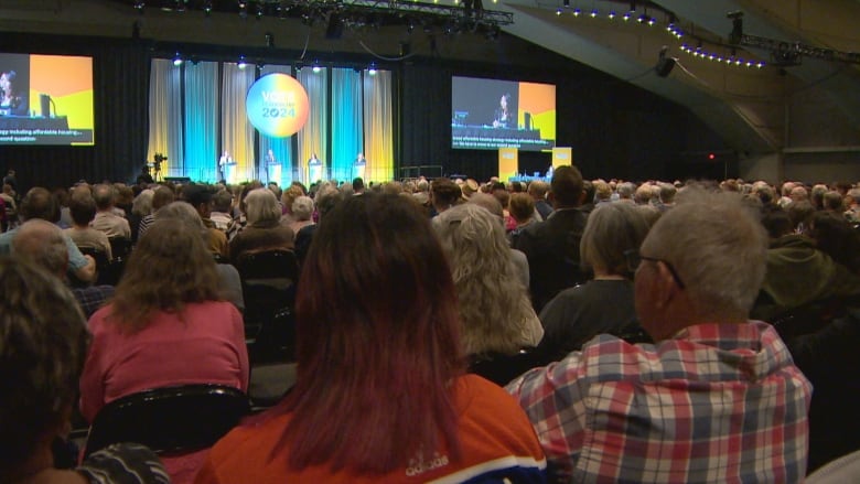 People sit inside a convention centre looking toward the stage.