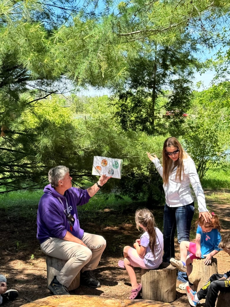 Two children sit on stumps as a man reads from a book. A woman has her hand on one of the children's heads. There are trees in the background.