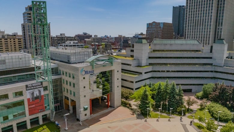 A photo taken with a drone of Ottawa City Hall downtown. The sky is sunny and the trees are green. The courtyard is relatively empty.